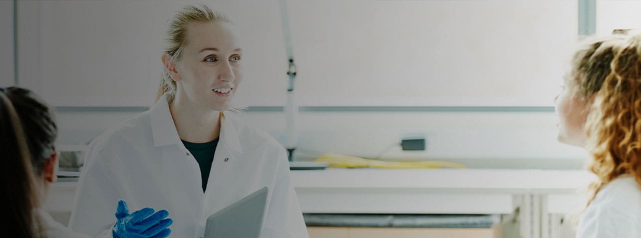 um cientista com uma bata branca de laboratório, luvas azuis e uma pasta, falando diante de um grupo de pessoas